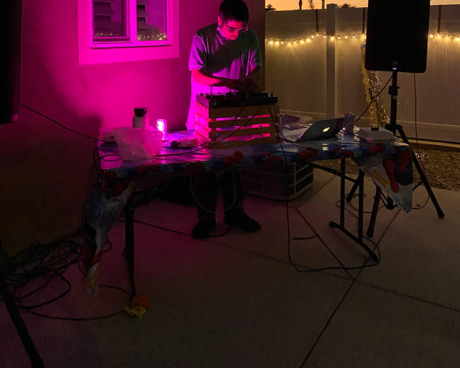 A person stands behind a table with equipment at a small outdoor event, illuminated by pink lights, against a sunset backdrop.