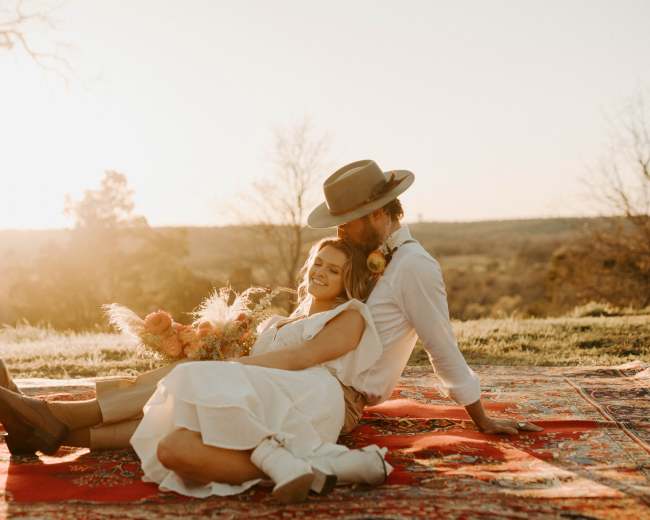 A couple sits on a patterned rug in a grassy landscape at sunset, with one person holding a bouquet of flowers.