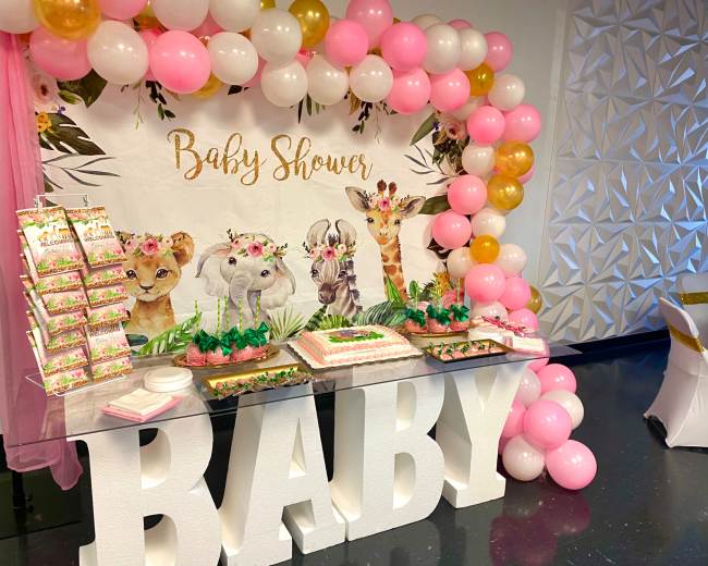 A decorated table for a baby shower featuring a cake, treats, and a backdrop with animal illustrations and balloons in pink and gold.