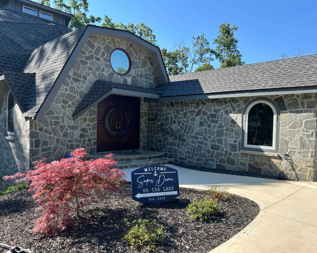The image shows a stone-faced house with a circular window and a welcoming sign in front, surrounded by landscaped greenery.