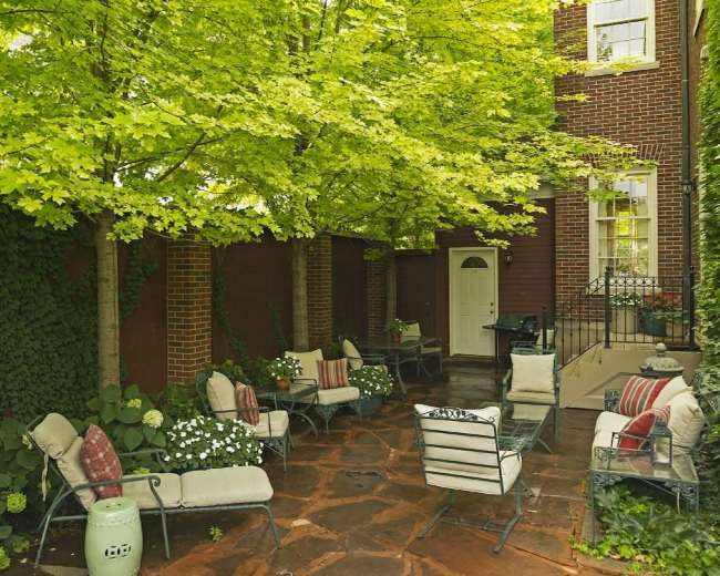 A patio area with stone flooring, surrounded by greenery, features several chairs and a door leading indoors.