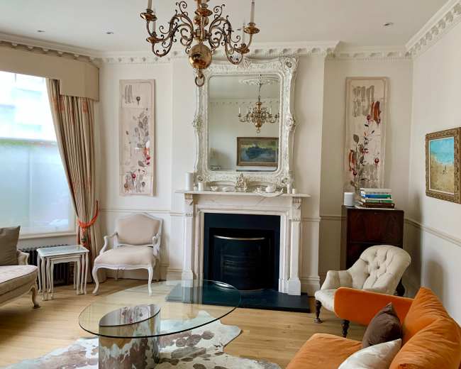 The image shows a living room featuring a mirror above a fireplace, a glass coffee table, an orange sofa, and decorative wall art.