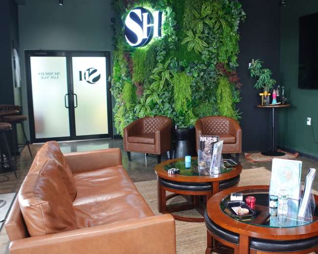 The image shows a modern lounge area with two brown leather chairs, round wooden tables, and a green wall covered in plants, alongside a large television and a bar setup.
