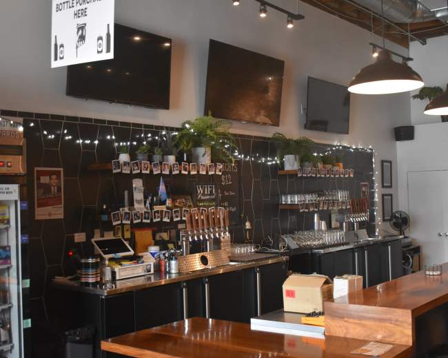 A bar area with a long wooden counter, multiple beer taps, a wall of framed photos, and several TVs mounted above the bar.