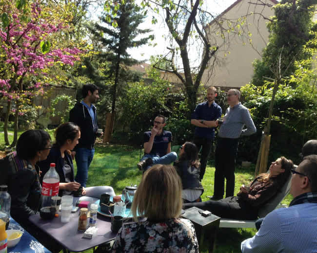 A group of people is gathered in a garden, engaging in conversation while seated and standing among greenery and blooming trees.