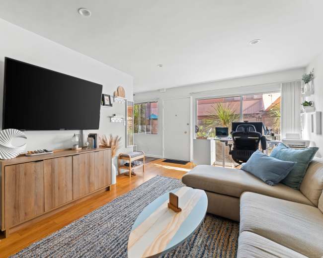 The living room features a large television on a wooden cabinet, a light-colored sectional sofa, and a coffee table, with natural light coming in through a window.