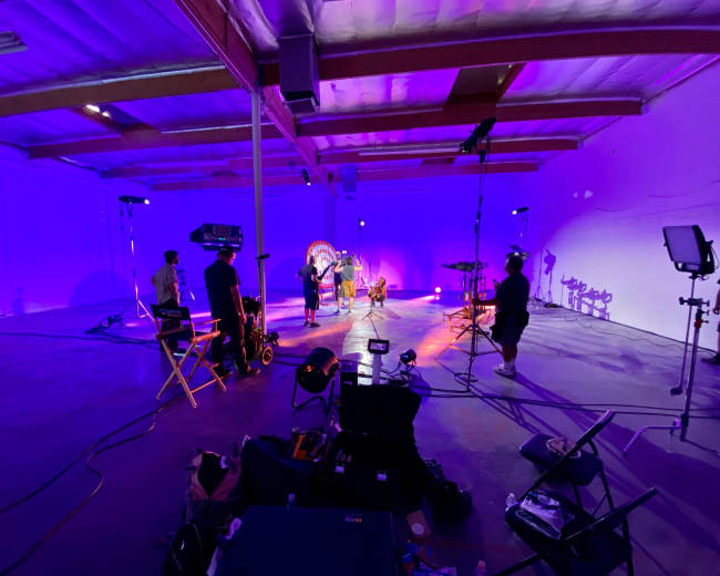 A group of people is setting up equipment and performing in a brightly lit, purple-toned studio space.