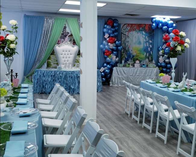 A decorated event space with tables set for a celebration, featuring white chairs, blue tablecloths, and colorful balloon arrangements.
