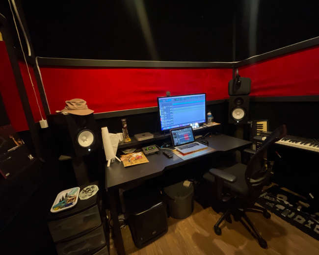The image shows a music production studio featuring a desk with a computer monitor, speakers, a keyboard, and various music production equipment against a backdrop of red and black walls.