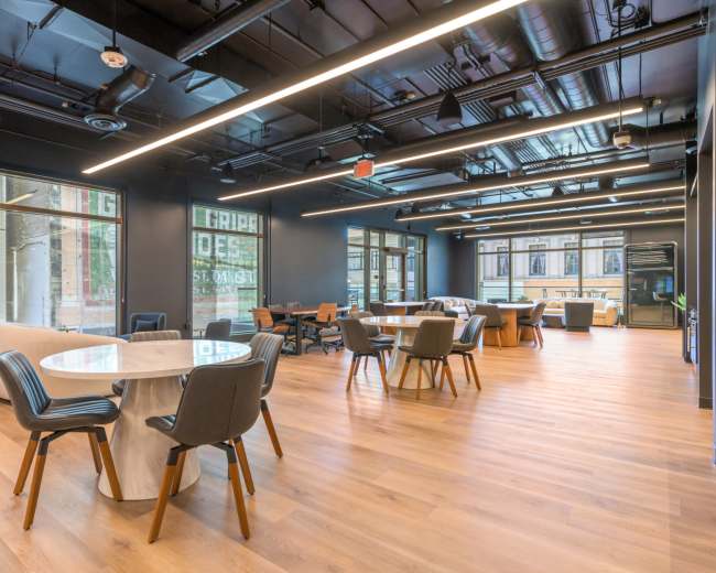 The image depicts a modern office space with wooden flooring, large windows, and various seating areas consisting of tables and chairs.