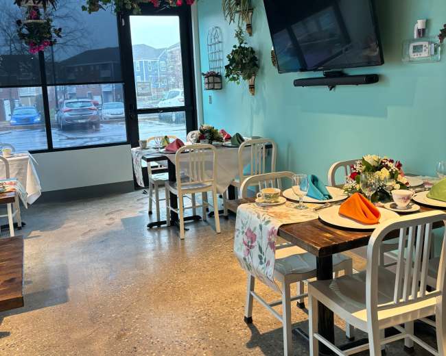 A brightly lit restaurant interior with wooden tables and white chairs, adorned with colorful table settings and hanging flower baskets.