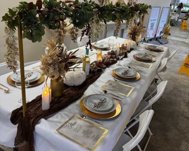 A long table is elegantly set with white tablecloths, gold accents, and a decorative arrangement of greenery and flowers for a special event.