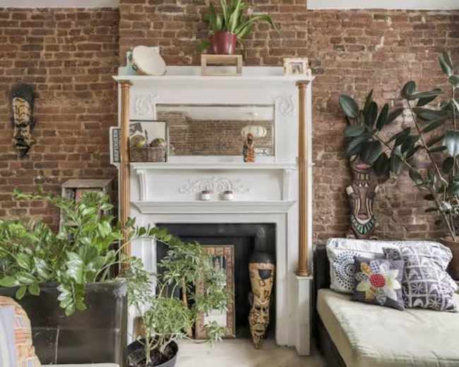 The image shows a cozy living space with a brick wall, a white fireplace mantel adorned with various decorative items, and plants in pots arranged nearby.