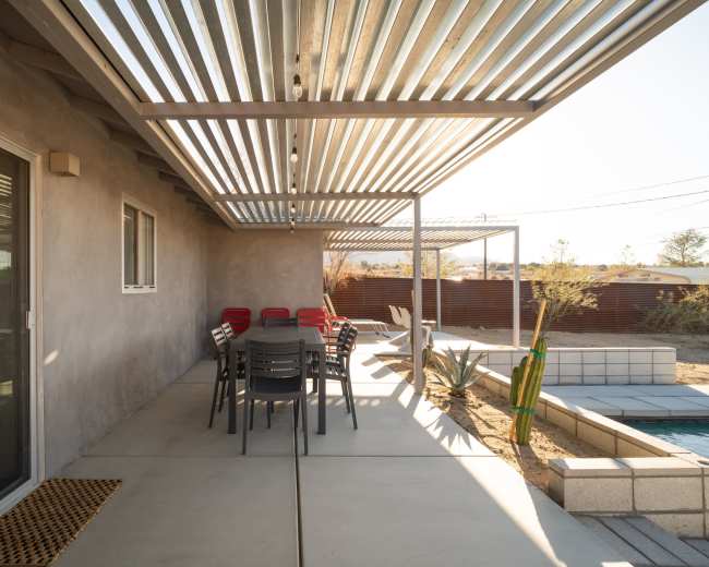 A concrete patio features a long dining table with chairs under a metal awning, surrounded by a landscaped yard with cacti and a pool.