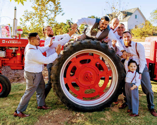 A group of men poses playfully around the large wheel of a red tractor in a grassy area.