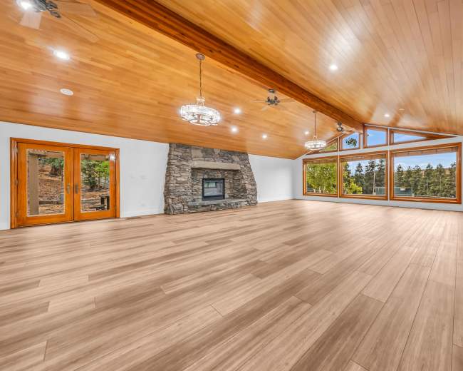 A spacious room features a stone fireplace, wooden ceiling beams, and large windows overlooking a wooded area.