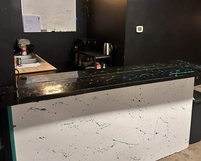The image shows a dark room with a white reception desk featuring a splatter design, a wooden countertop behind it, a whiteboard on the wall, and some greenery decor.