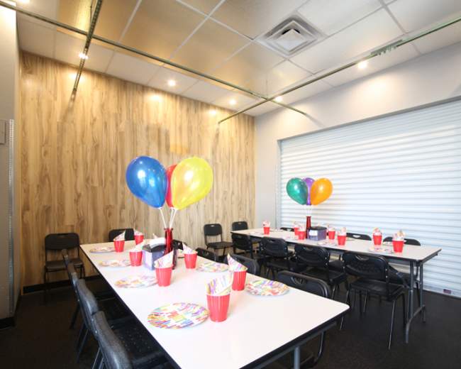 A decorated party room features a long table set with colorful plates and cups, surrounded by chairs and adorned with balloons.