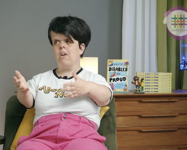 A person with short hair is seated on a green chair, wearing a white shirt and pink pants, while gesturing with their hands and discussing books displayed on a nearby shelf.