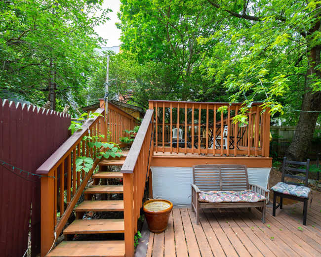 A wooden deck with chairs and a staircase leads to a patio surrounded by lush green trees.