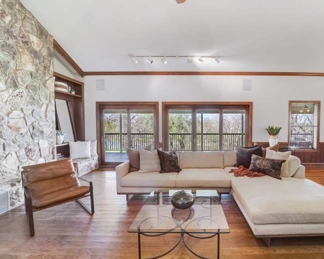 A spacious living room features a large sectional sofa, a glass coffee table, and a stone fireplace, with large windows offering views of the outdoors.