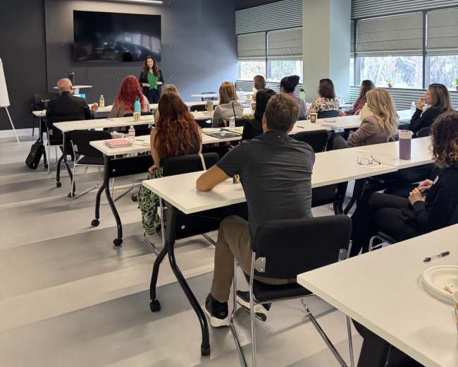 A group of attendees sits in a seminar room, facing a speaker who is presenting near a large screen.