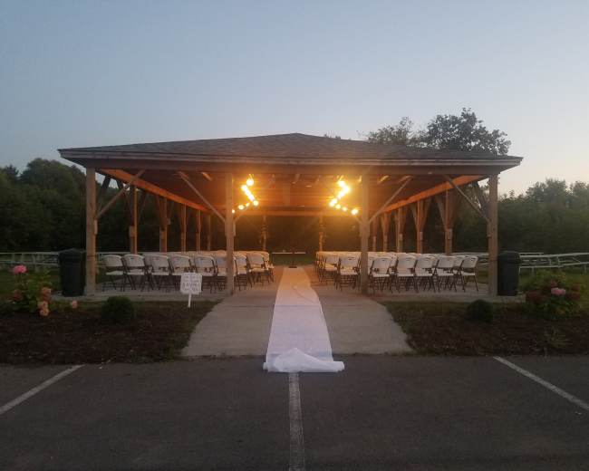 A covered pavilion is set up with rows of white chairs and a white carpet leading to the center, illuminated by overhead lights, surrounded by greenery.