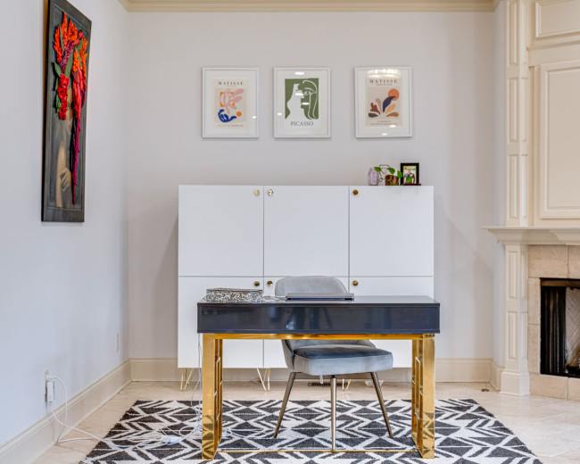 The image shows a modern home office with a sleek black desk and a chair, a white cabinet, framed artwork on the walls, and a fireplace with a marble surround.