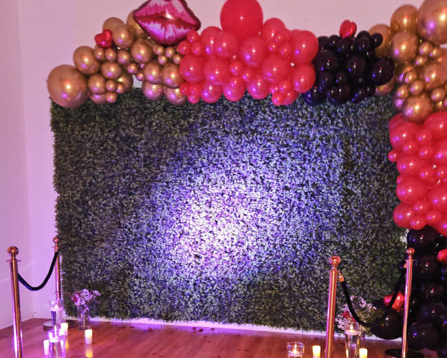 A decorated event space with a wall of greenery adorned with red, gold, and black balloons, and a red carpet entrance marked by stanchions and rose petals scattered on the floor.