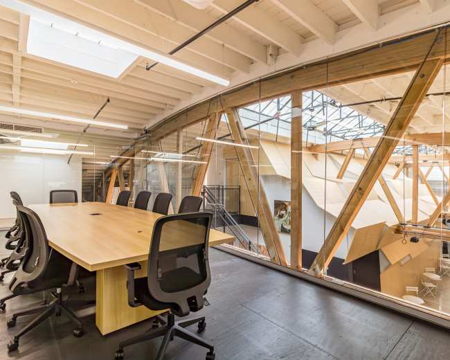 The image shows a modern conference room with a large wooden table surrounded by black chairs, situated within an open office space featuring exposed wooden beams and large glass walls.
