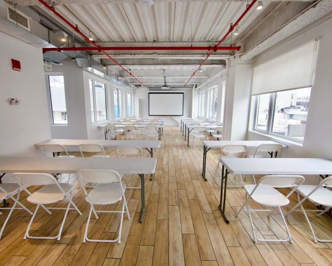 A bright, empty meeting room with rows of tables and chairs arranged in a grid, a projector screen at the front, and large windows letting in natural light.