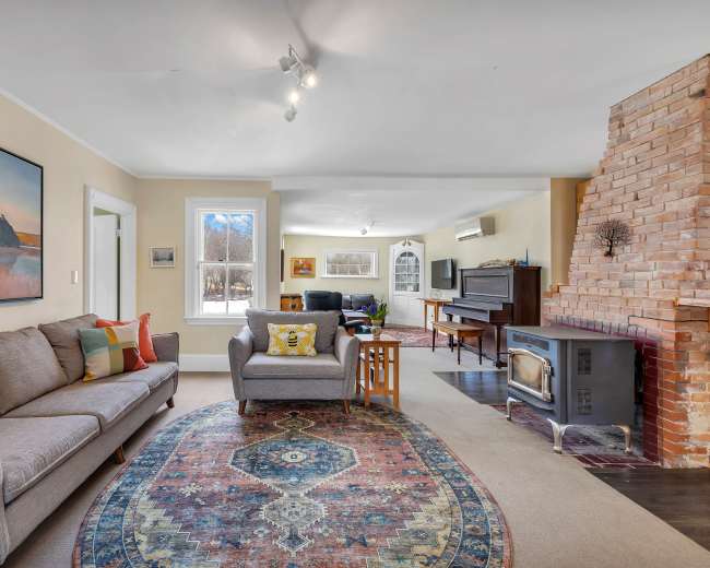 A spacious living room features a cozy sofa, a patterned rug, a wood-burning stove, and a piano, with large windows allowing natural light to enter.