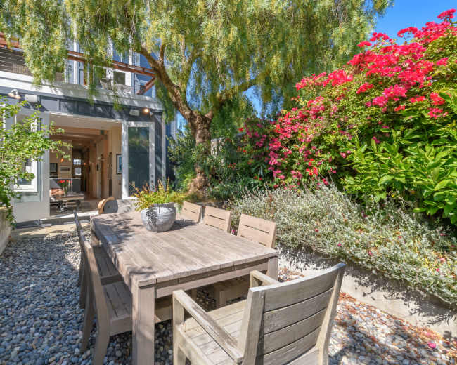 A wooden dining table with chairs is set on a gravel patio, surrounded by green plants and vibrant flowering bushes, near a house with a large open doorway.