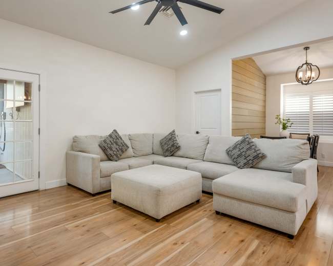 The image shows a light-colored sectional sofa with decorative pillows arranged in a living room with wooden flooring and a door leading outside.