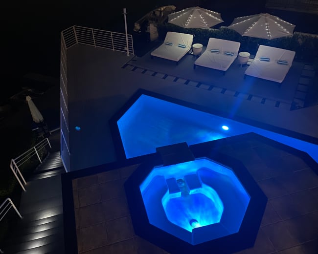 The image shows a modern rooftop area featuring a swimming pool and a hot tub, illuminated by blue lights, with loungers and umbrellas nearby.