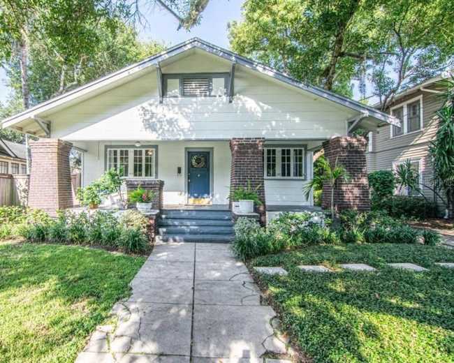 A single-story house with a white exterior and a blue front door is flanked by brick columns and surrounded by a well-maintained lawn and garden.
