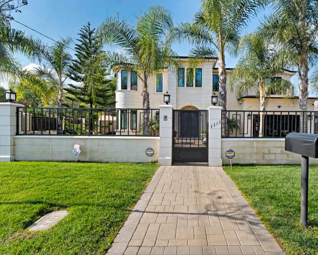 A modern two-story house with large windows is surrounded by a black metal fence and palm trees, with a paved walkway leading to the front gate.
