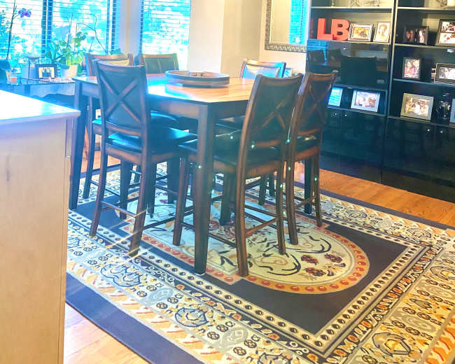 A dining area features a wooden table surrounded by chairs, a modern light fixture above, and large windows with blinds, alongside a black bookshelf displaying framed photos.