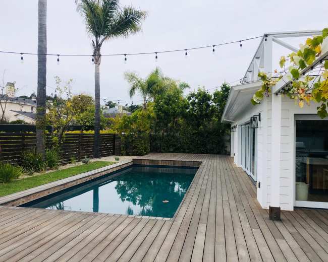 A wooden deck surrounding a rectangular swimming pool, with palm trees and string lights visible in a landscaped backyard.