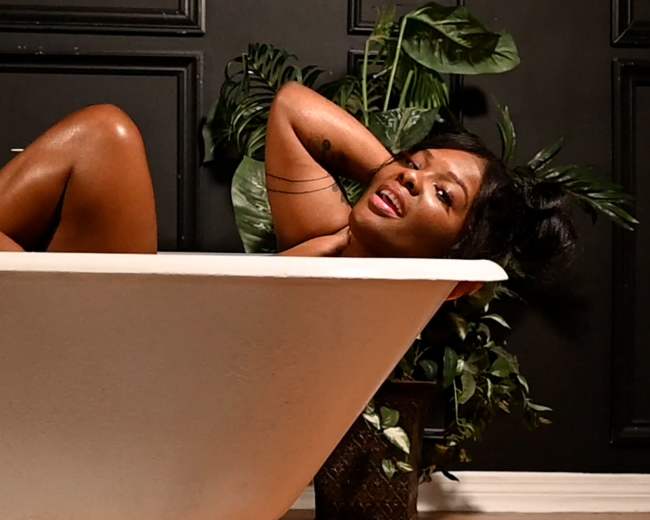 A person is partially submerged in a freestanding bathtub surrounded by indoor plants.