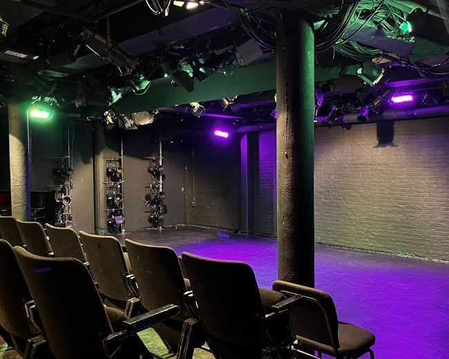 The image shows a small theater stage with rows of empty seats facing a dimly lit performance area, illuminated by colored stage lights.