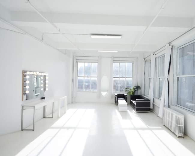 The image shows a bright, minimalist studio space with large windows, a mirror with lights, a plant, and modern furniture.