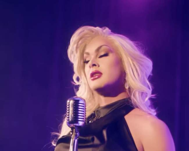 A performer stands in front of a vintage microphone, dressed in a sleek black outfit with styled blonde hair, set against a purple-lit backdrop.