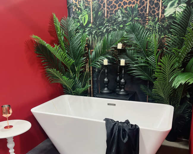 The image features a modern white bathtub with a black drape, surrounded by lush green plants against a red wall and a decorative backdrop.