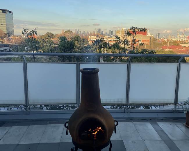 A chiminea with a fire is positioned on a balcony overlooking a city skyline and landscaped greenery.