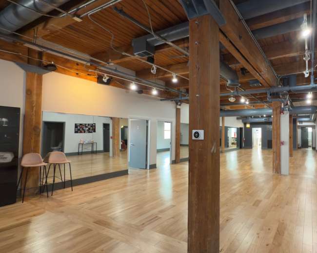 The image shows an empty, spacious room with wooden floors, exposed beams, and large mirrors along one wall.