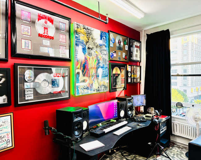 The image shows a music production studio featuring a black desk with computer monitors, music equipment, and a colorful wall adorned with framed records and art.