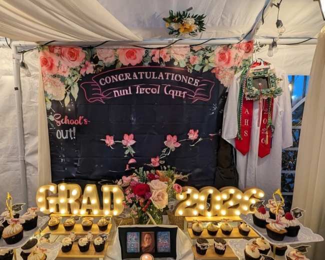 The image shows a decorated table with a graduation-themed backdrop featuring cupcakes arranged on white platters, lit "GRAD" and "2023" signs, and a celebratory banner that reads "CONGRATULATIONS."