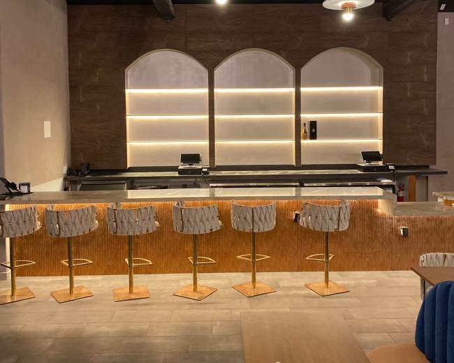 The image shows a modern bar area with arched wall features, illuminated shelves, and upholstered stools arranged along the counter.