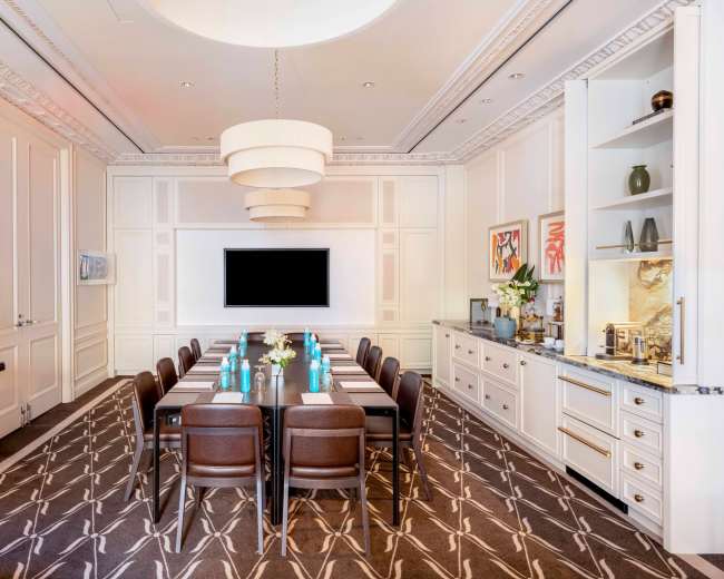 The image shows a modern conference room featuring a long table with ten brown chairs, a large wall-mounted screen, and elegant decor including a chandelier and side cabinet.